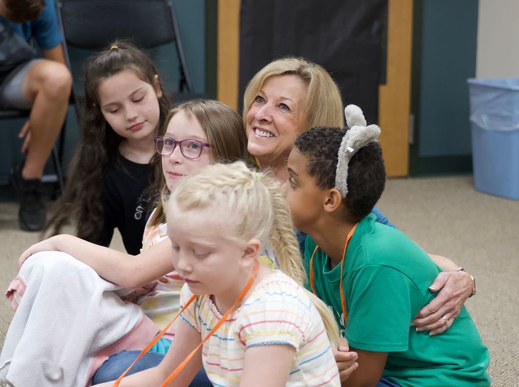 A group of children are sitting around a woman with bunny ears on her head.
