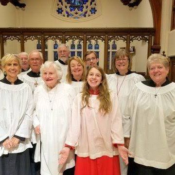 Choir in church: diverse group in white robes and pink/red attire, singing in a wooden interior.
