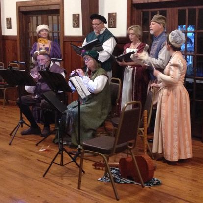 Musicians in Medieval costume perform with music stands on a wooden floor.