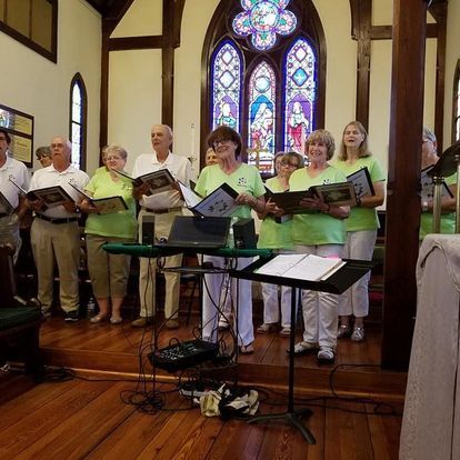 Choir singing in a church. People in green shirts, sheet music, stained glass window.