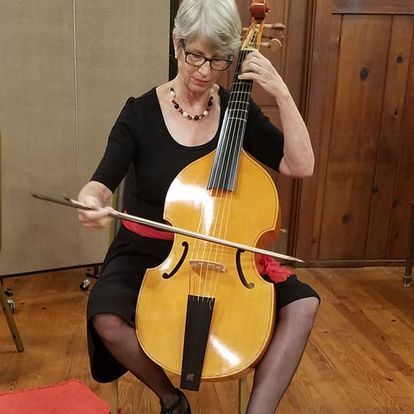 Woman playing a viola da gamba, seated indoors; blonde hair, black dress, red sash, holding bow.