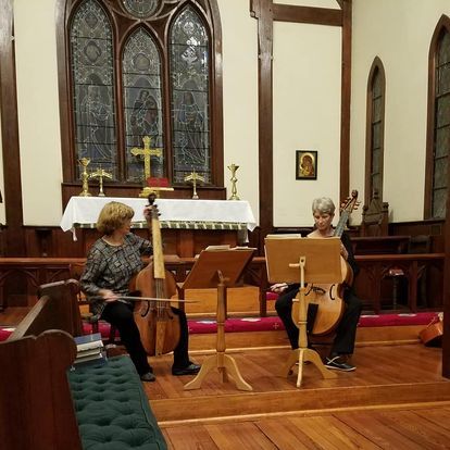 Two women playing cellos in a church. One with dark hair, other with short silver hair. Wooden floors and pews.