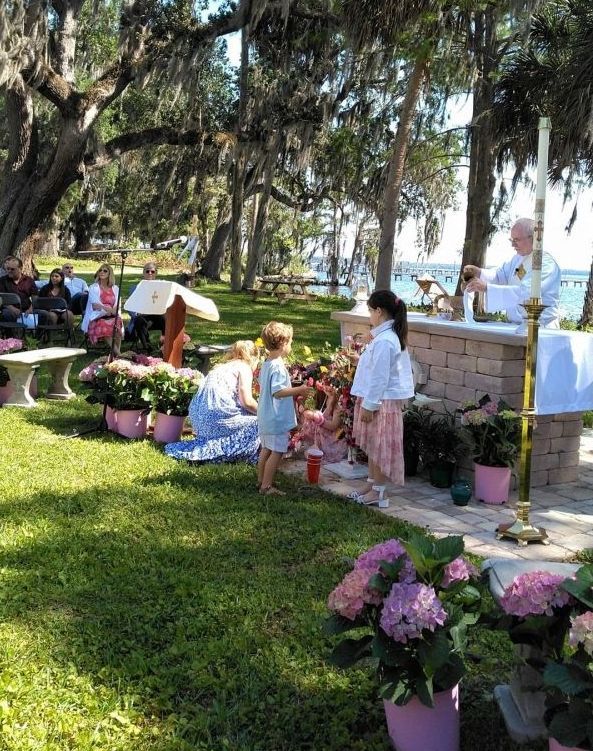 Outdoor religious ceremony with children offering flowers; clergy at altar, pink hydrangeas, water backdrop.