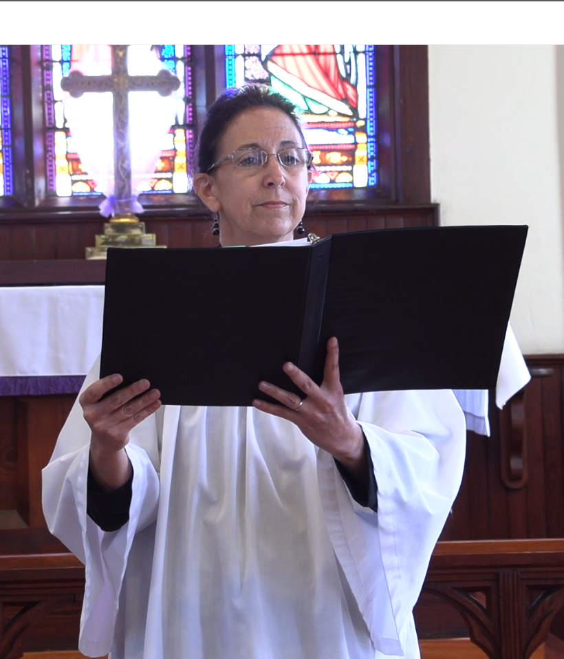 Cantor in white robe, glasses, reading from a black book in church. Stained glass and a cross in the background.