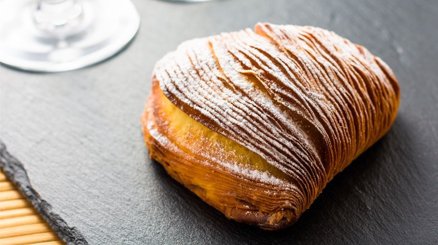 Un uomo in camice nero da chef sorride, tenendo in mano una grande palla bianca, vicino a una scatola. Bar con bottiglie.