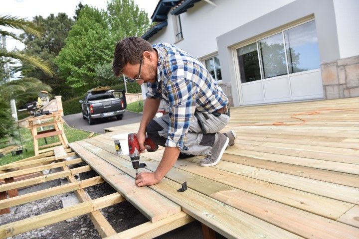 A person wearing a plaid shirt kneels on a wooden deck frame, using a drill to secure a board to the joists outdoors.