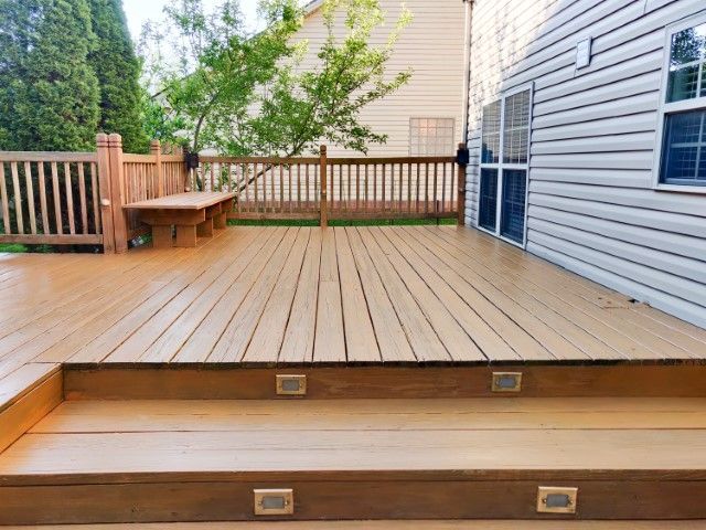 A two-level wooden deck with built-in stairs, recessed step lighting, and a railing overlooking a backyard with trees.