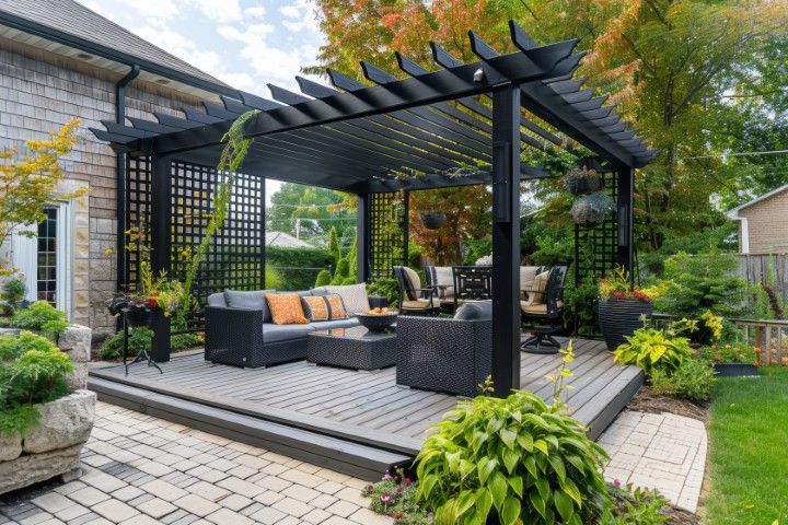 A black pergola stands over a grey wooden deck with modern outdoor furniture in a lush, landscaped backyard.