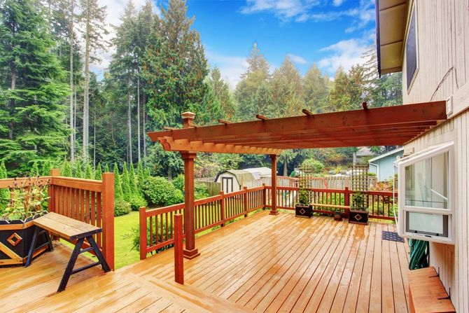 A wooden deck with a pergola attached to a house, overlooking a lush backyard surrounded by tall pine trees.
