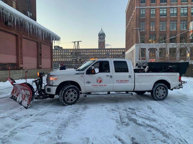 White pickup with red snowplow parked on snowy street in front of buildings and a clock tower