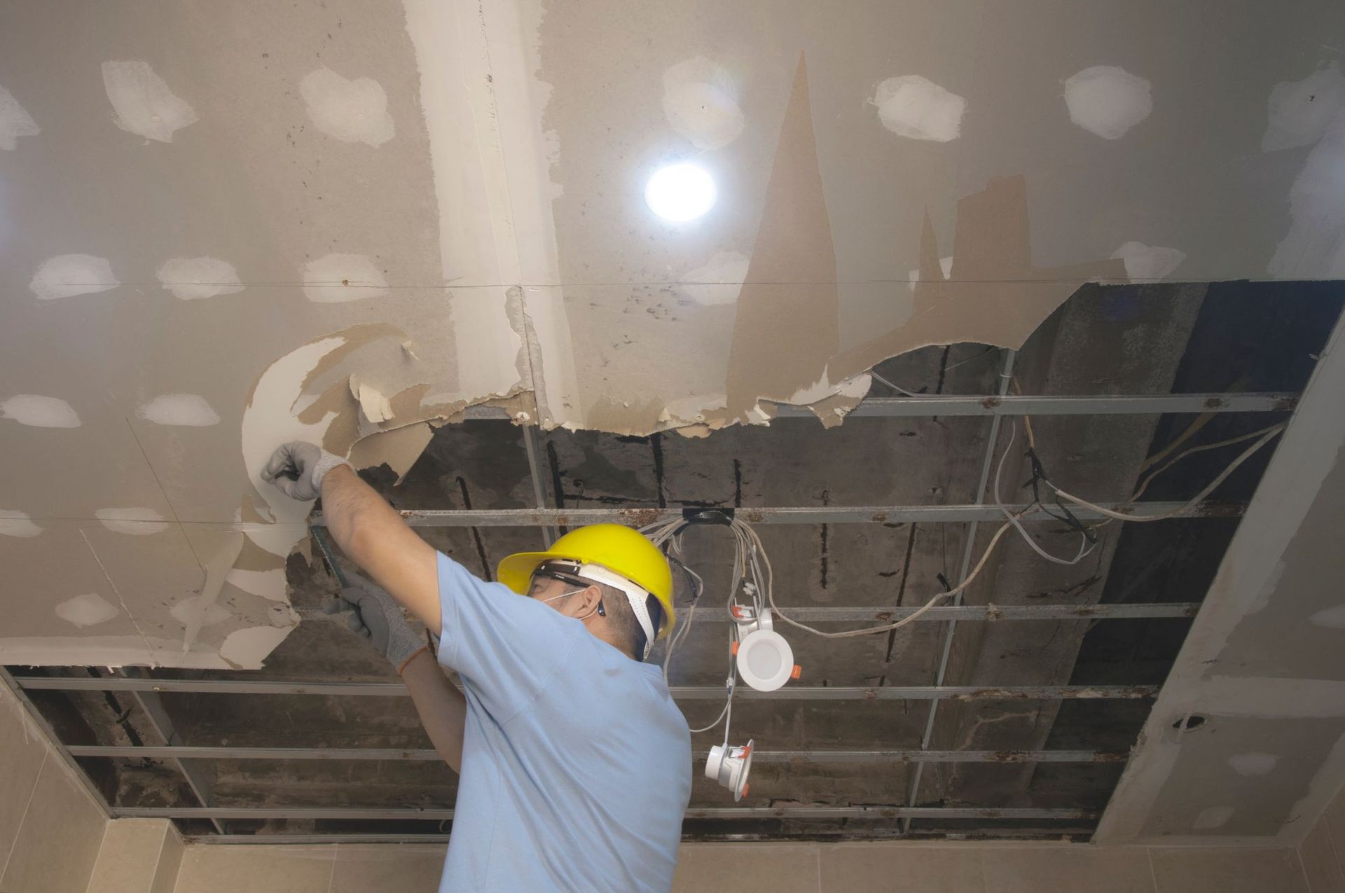Worker in a hard hat removing drywall from a damaged ceiling with exposed wiring.