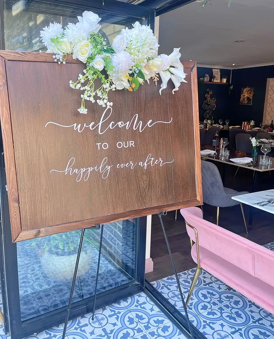 A wooden sign with flowers on it is sitting in front of a restaurant.