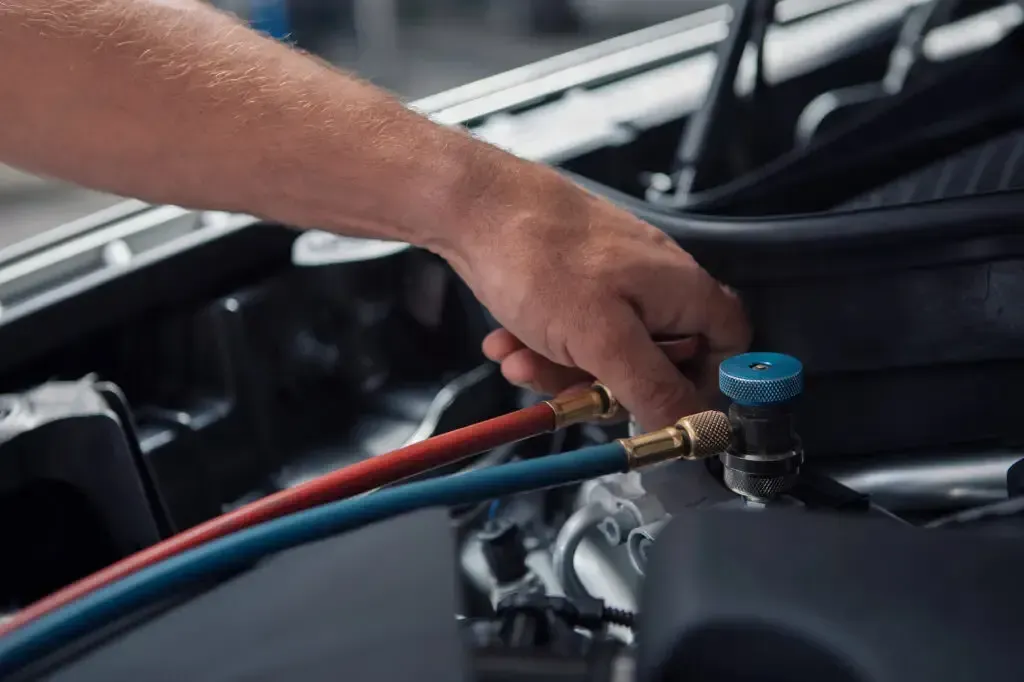 A hand connects blue and red hoses to an air conditioning service port under the hood of a car.