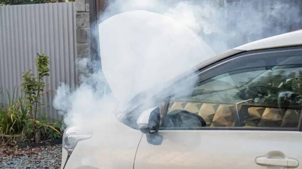 A white car parked outdoors with its hood open, emitting a thick cloud of white smoke from the engine bay.