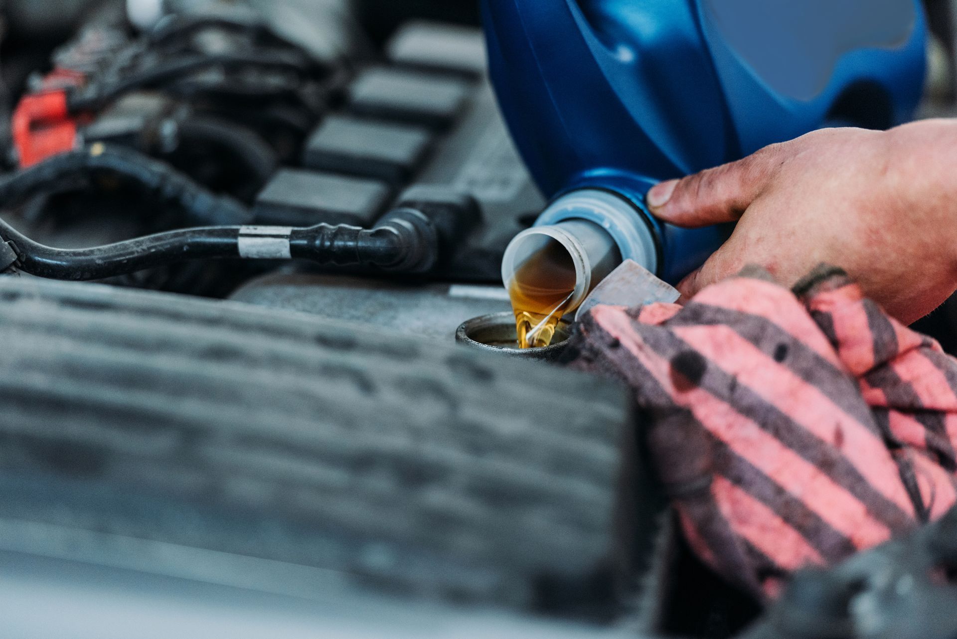 A hand in a striped work glove pours golden motor oil from a blue plastic bottle into an engine.