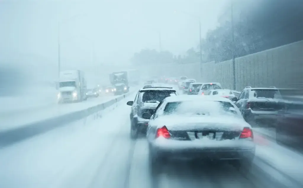 Traffic moves slowly on a highway during a heavy snowstorm, with snow-covered vehicles driving in low visibility.