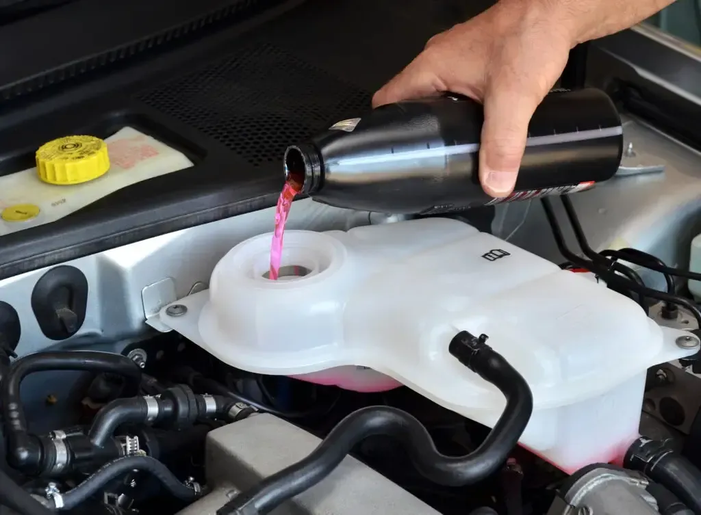 A person pouring pink coolant from a black bottle into the reservoir of a car engine.