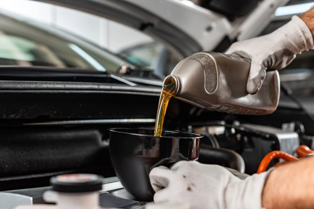 Hands in white gloves pouring golden motor oil into a funnel inside an open car hood.