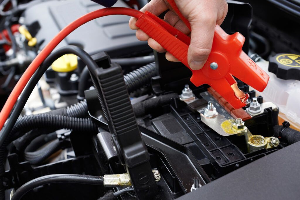 A hand connecting red jumper cables to a car battery terminal in an engine bay.