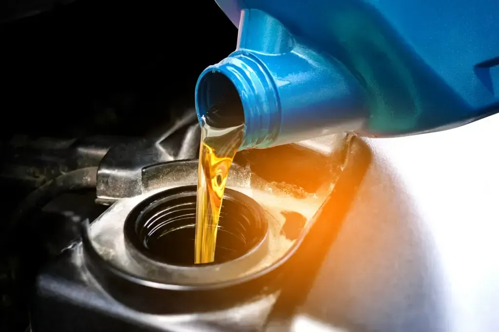 Golden motor oil pouring from a blue plastic bottle into a car engine oil filler neck.