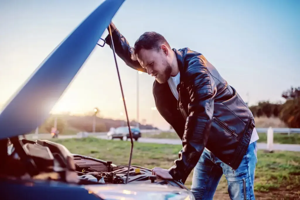 A person in a leather jacket looks frustrated while inspecting the open hood of a broken-down car at sunset.