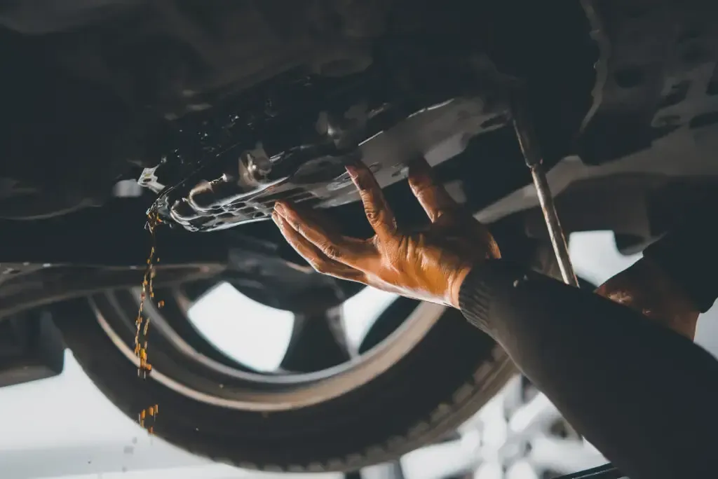 A mechanic performs an oil change, with used oil draining from the underside of a car into a container.
