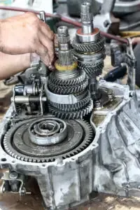 A mechanic's hands work on the complex internal gears and shafts of an exposed automotive manual transmission.