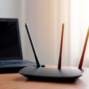 Black Wi-Fi router with three antennas, near a laptop on a wooden surface, with a bright window in the background.
