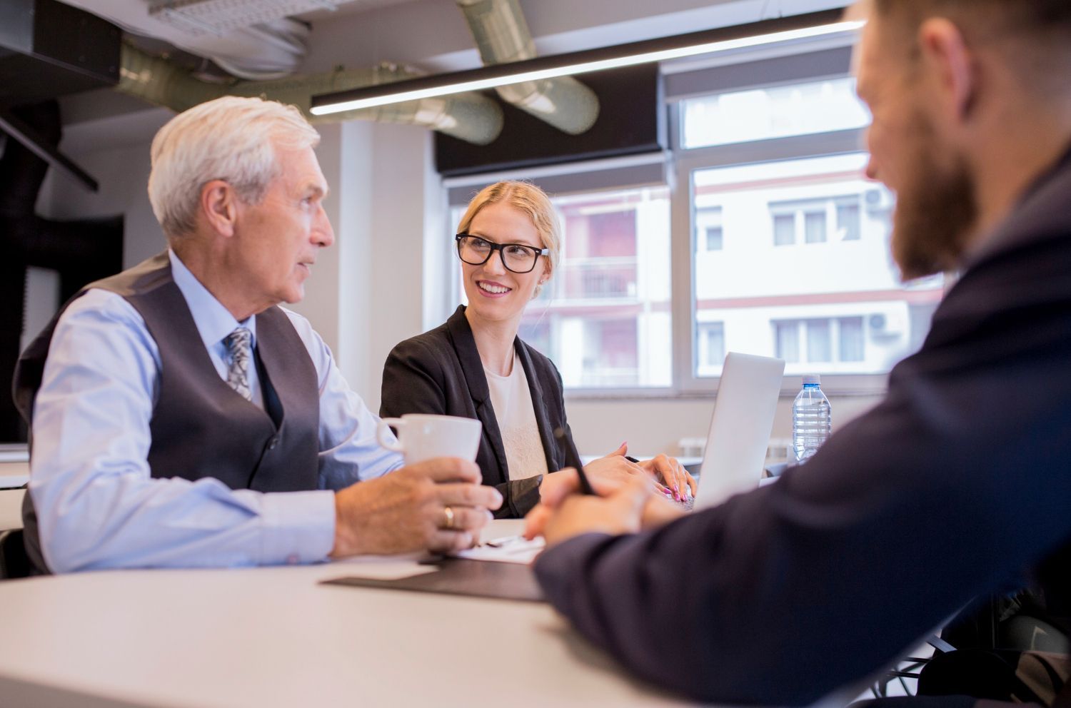 Three colleagues discussing around a conference table in a bright office, with a laptop and coffee cups.