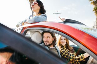 Two people inside a red car with another person standing through the sunroof, all smiling outdoors
