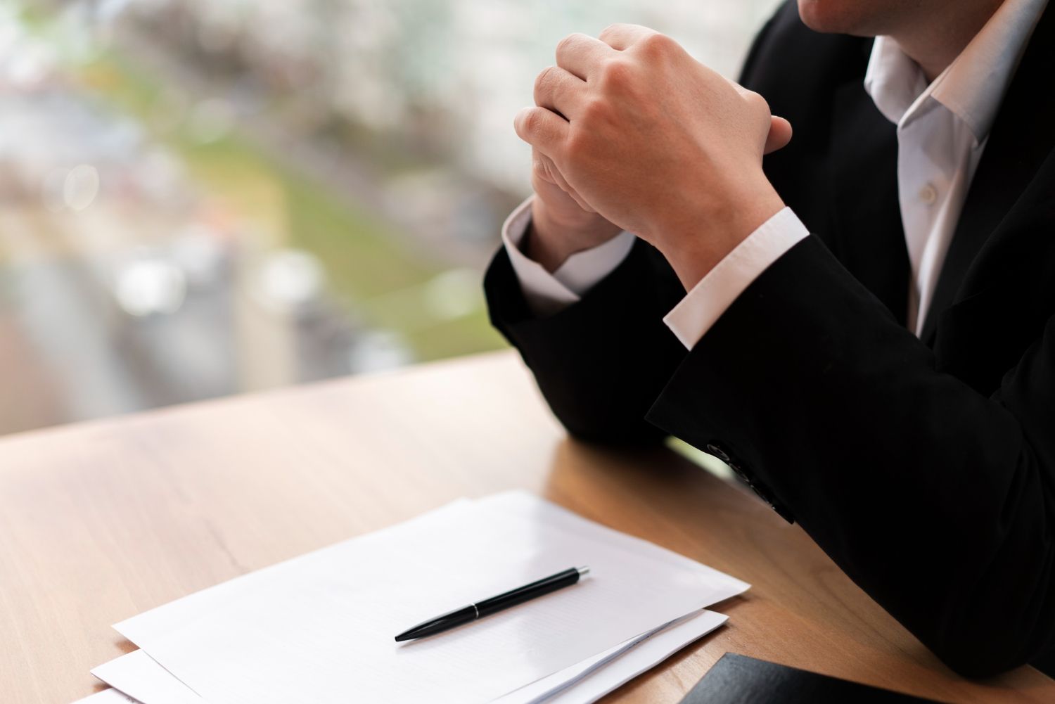 Person in a black suit sits at a table with a notebook and pen, hands clasped thoughtfully.