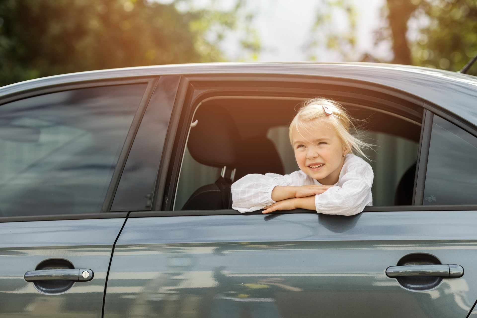 Smiling child leaning out of a car window, resting on the door in daylight