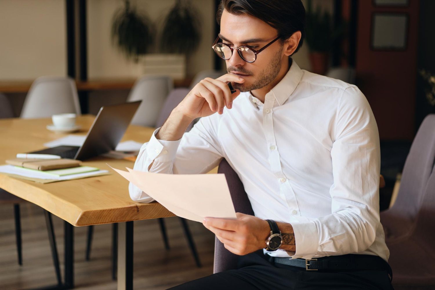 Man in a white shirt reviewing a document at a desk with a laptop in a modern office