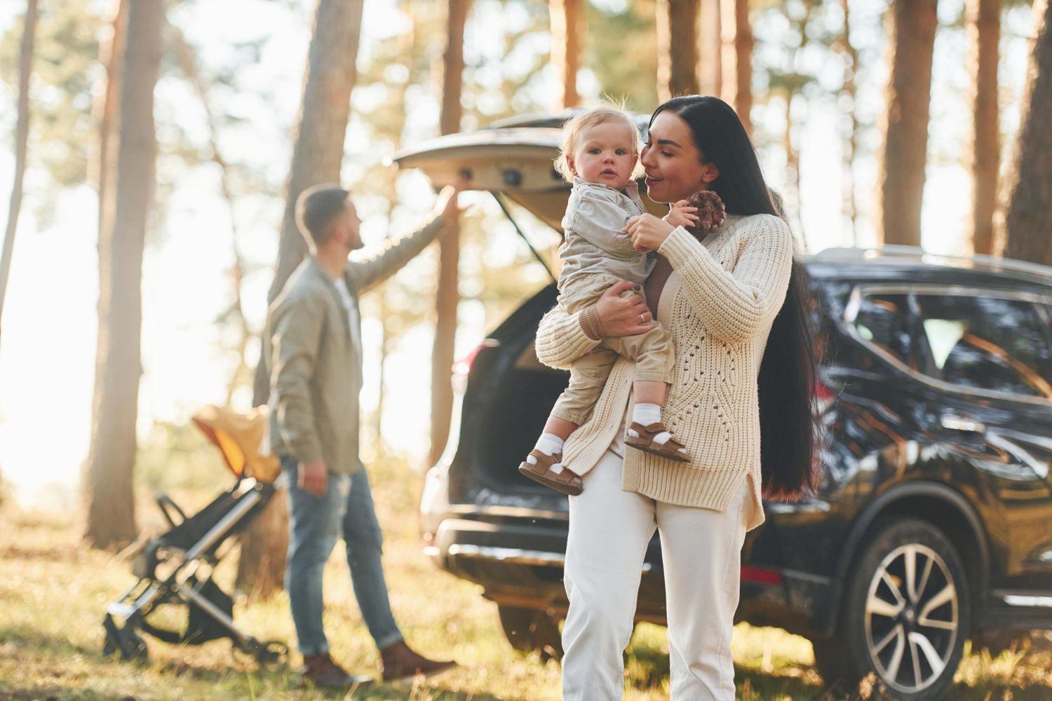 Woman holding a child beside an SUV in a sunlit forest campsite, with a stroller nearby