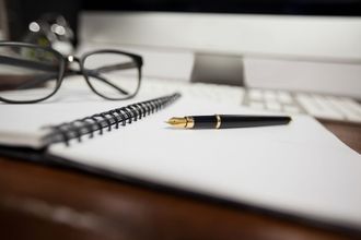 Blank notebook with a black fountain pen on a desk, with eyeglasses in the background
