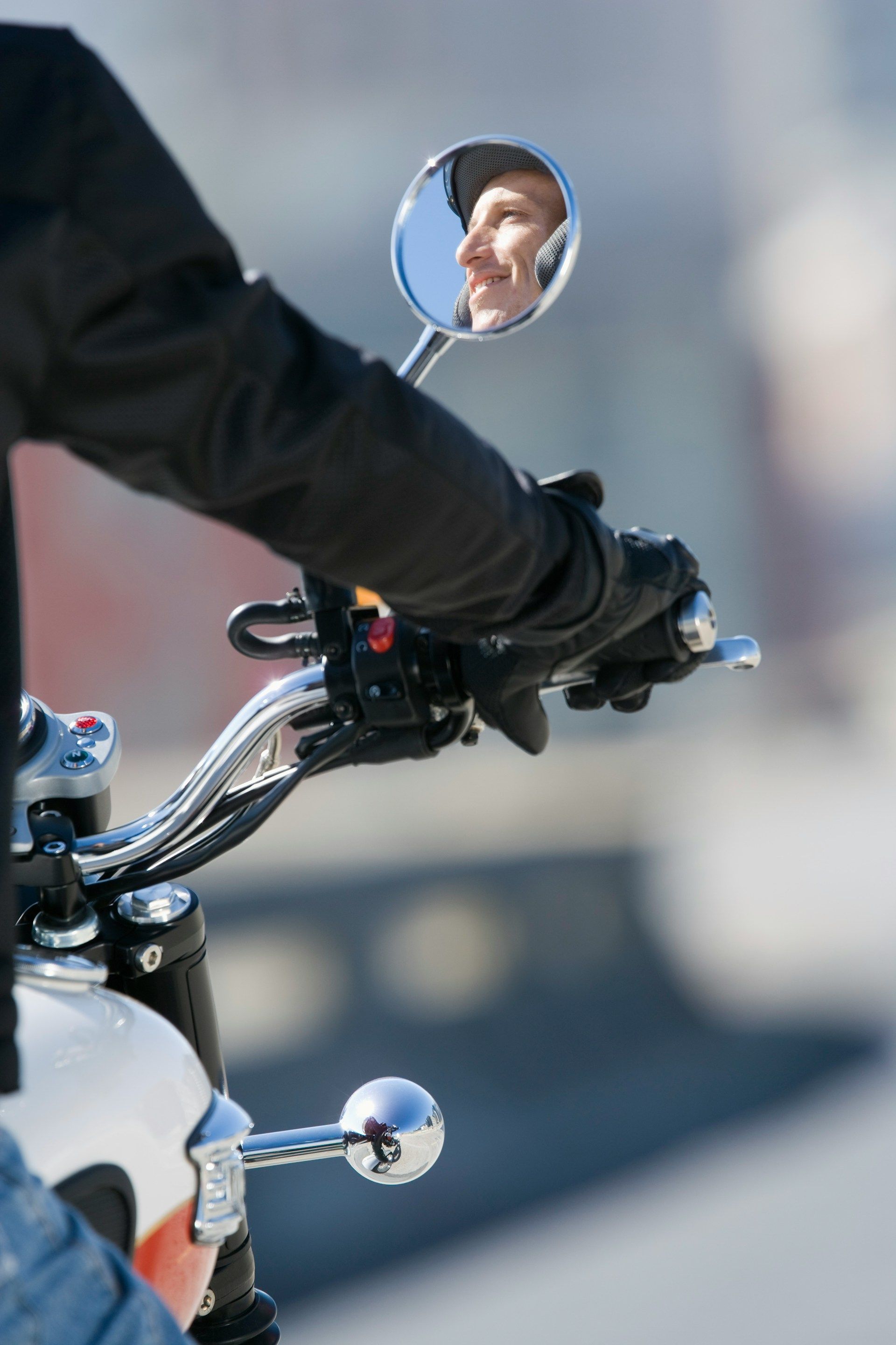 Motorcycle handlebar with rider’s gloved hand and rearview mirror reflecting a face outdoors