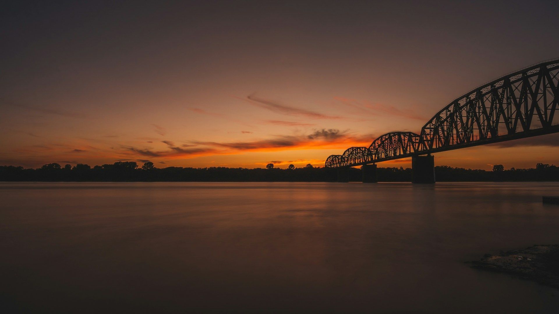 Sunset over calm water with a silhouetted steel bridge stretching across the horizon
