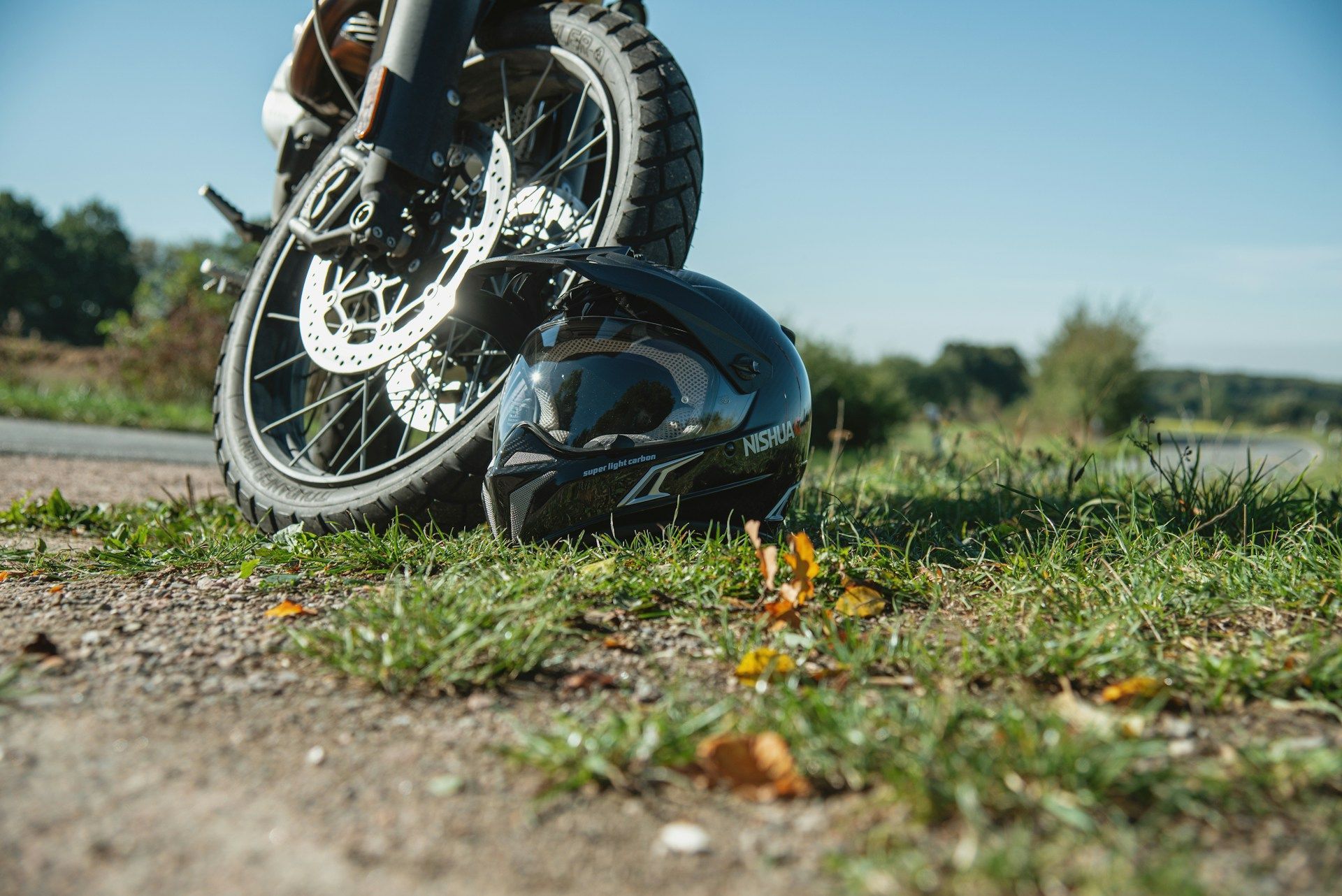 Motorcycle wheel crushing a glossy black helmet on a dirt road, with grass and trees in the background