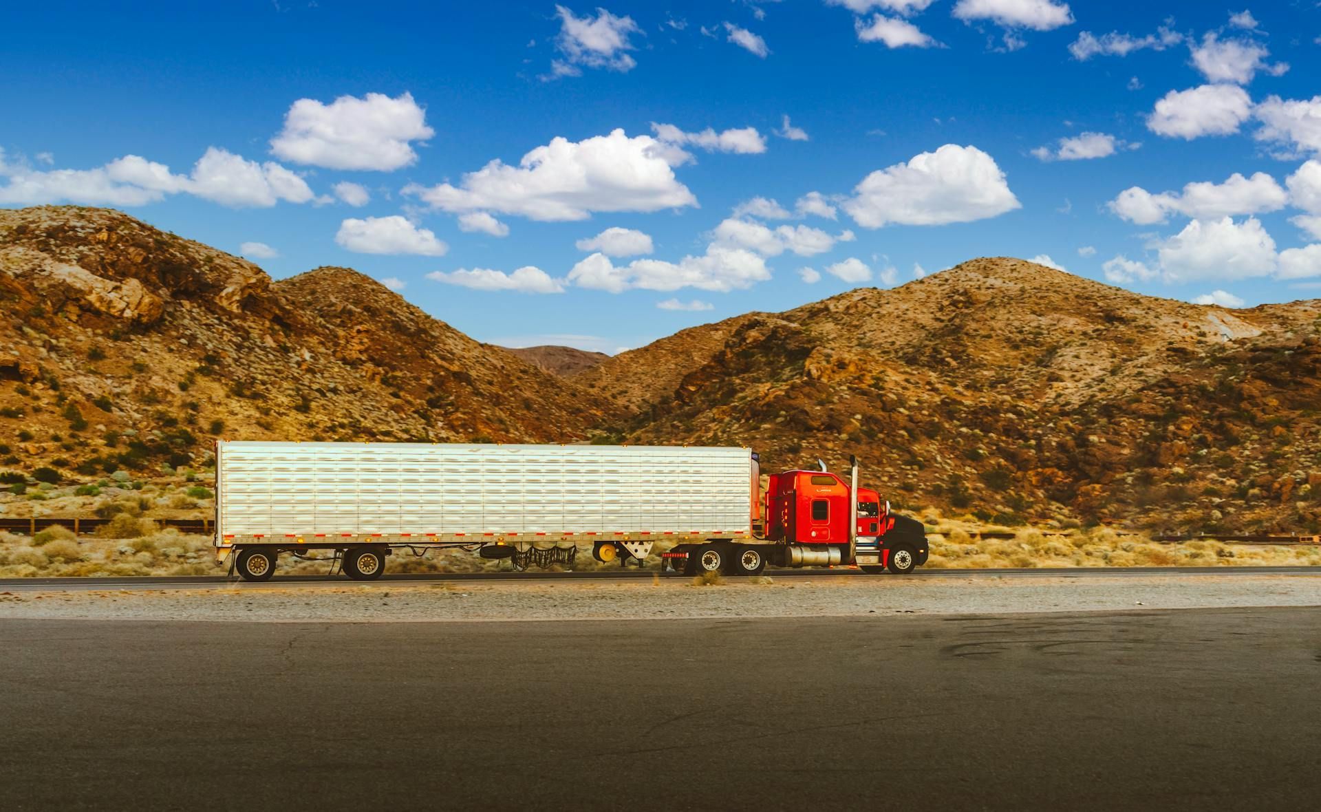 Red semi-truck hauling a white trailer across a desert road with rocky hills under a blue sky