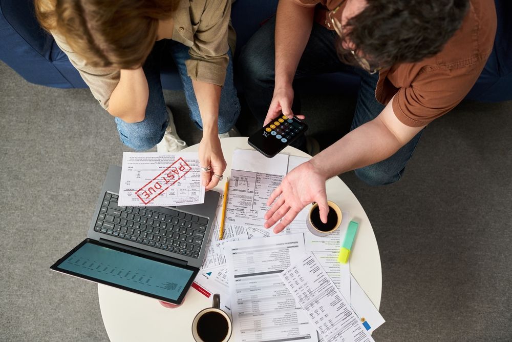 Three people review papers around a laptop and coffee mug at a table with highlighters and a calculator.