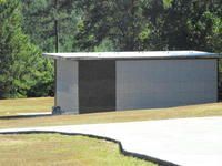 Small gray utility building beside a paved area, with trees and grass in the background.