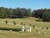Grassy park landscape with a pond, trees, and small white statues along a path under a blue sky