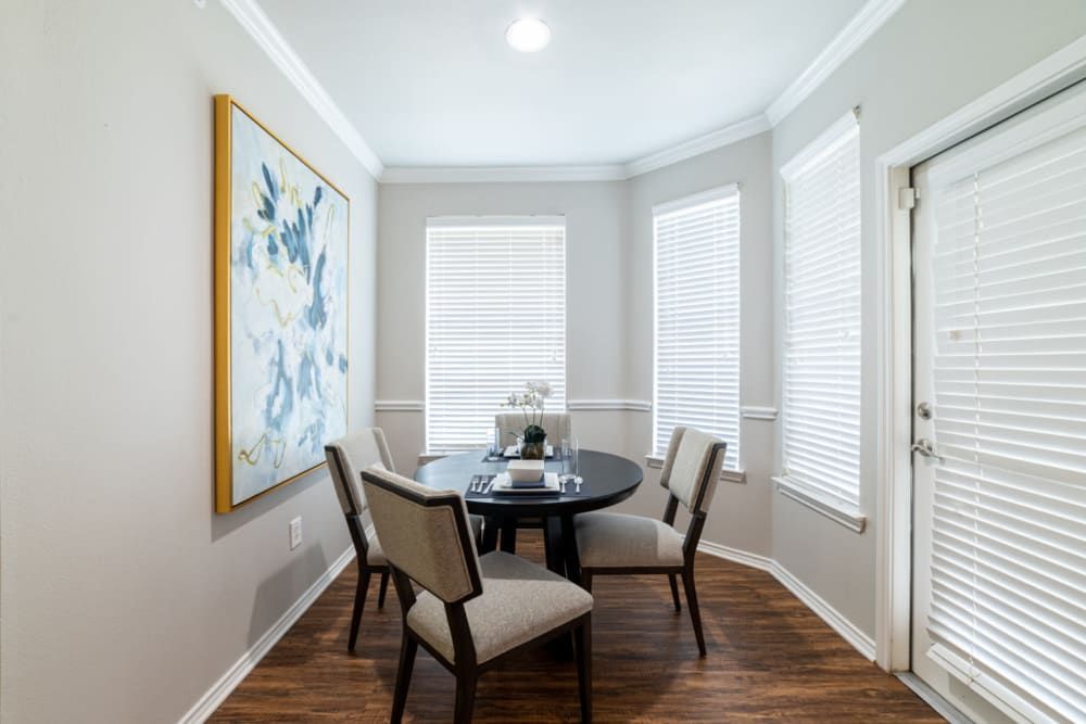 A well-lit dining room with a round black table and four upholstered chairs, large windows with white blinds, and a large abstract blue and white painting on the wall at Marquis at Bellaire Ranch in Fort Worth, TX.