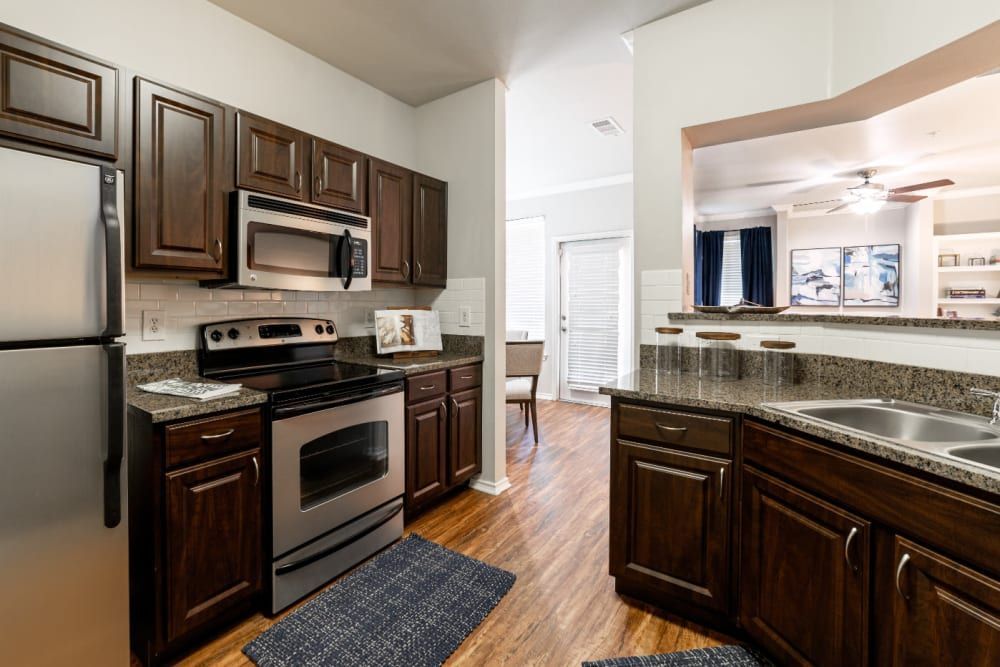 Modern kitchen interior with dark wood cabinetry, stainless steel appliances, and granite countertops. A kitchen island with a double sink faces an open concept dining area with a large window dressed in blue curtains. The floor is covered with hardwood, and a patterned blue rug lies in front of the stove at Marquis at Bellaire Ranch in Fort Worth, TX.