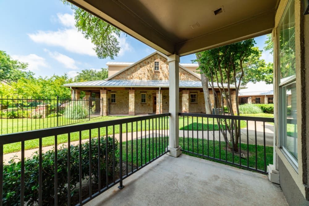View from a shaded porch overlooking a residential area, featuring a metal railing in the foreground with a stone house and landscaped garden in the background. Young trees and a well-maintained lawn are visible within the fenced property under a clear blue sky at Marquis at Bellaire Ranch in Fort Worth, TX.
