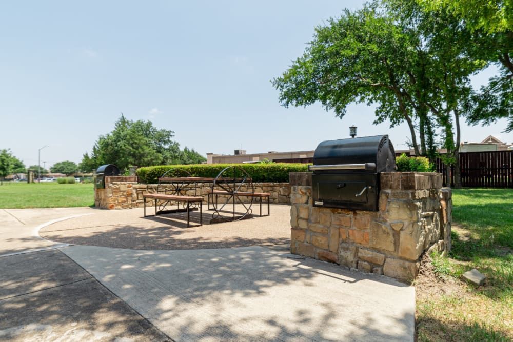 Outdoor barbecue area with stone-built grill station and picnic table set under the shade of vibrant green trees on a sunny day at Marquis at Bellaire Ranch in Fort Worth, TX.