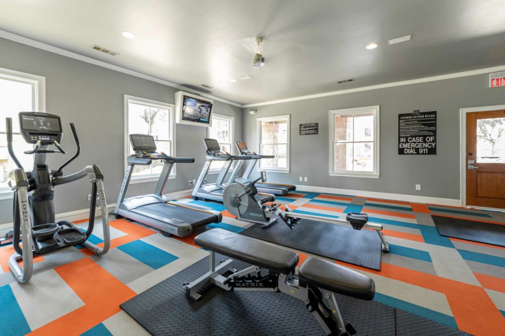 An indoor fitness gym with a variety of equipment including treadmills, an elliptical machine, and weight benches on a multicolored geometric-patterned floor. The room has grey walls adorned with fitness-related posters, and natural light is coming through the windows at Marquis at Bellaire Ranch in Fort Worth, TX.
