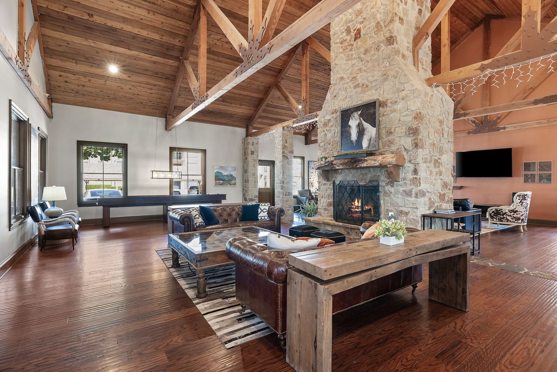 Rustic living room with vaulted wood ceiling, stone fireplace, wooden table, and large windows at Marquis at Bellaire Ranch in Fort Worth, TX