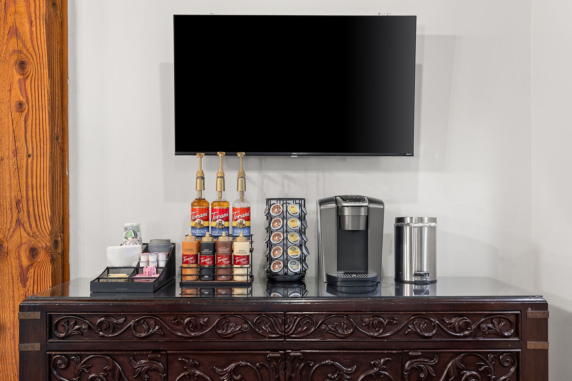 Wall-mounted TV above a carved wooden cabinet with coffee setup and stacked cups in a lounge area at Marquis at Bellaire Ranch in Fort Worth, TX