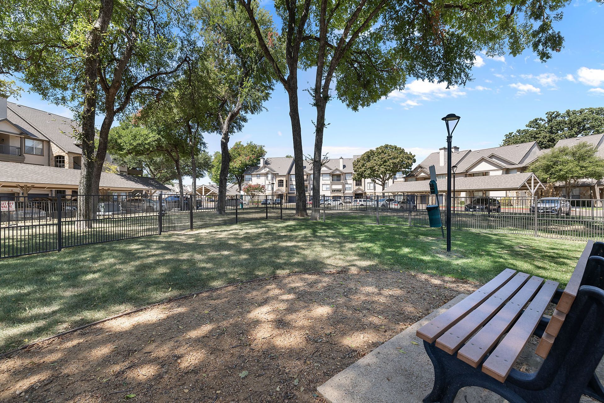 Park with trees, benches, and apartment buildings around a grassy open area under a clear sky at Marquis at Bellaire Ranch in Fort Worth, TX.